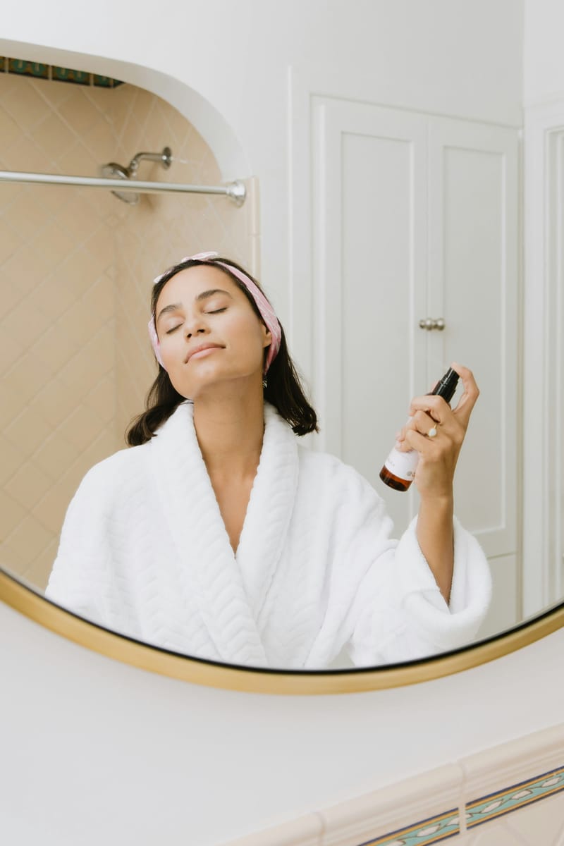 Femme en peignoir vaporisant une brume sur son visage devant un miroir.